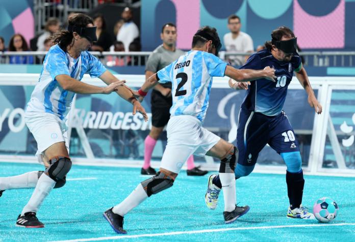 Two blind football players in Argentina uniforms and black eyeshades chase a French player, who has the ball