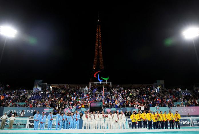 Three blind football teams stand on a podium, with the Eiffel Tower in the background, Argentina in blue with the silver medal, France in white with the gold medal, and Brazil in yellow with the bronze medal