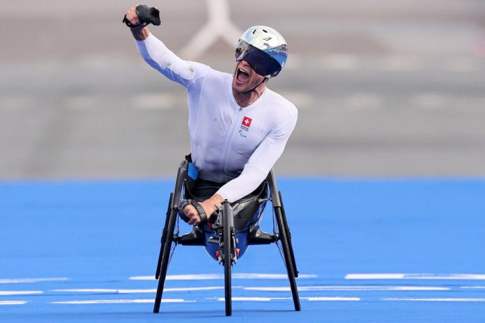 Man in a silver helmet, white outfit and racing wheelchair raises his right hand in the air and shouts