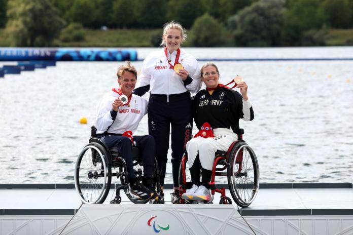 Tres mujeres, dos en silla de ruedas, sostienen medallas mientras están en un podio junto al agua