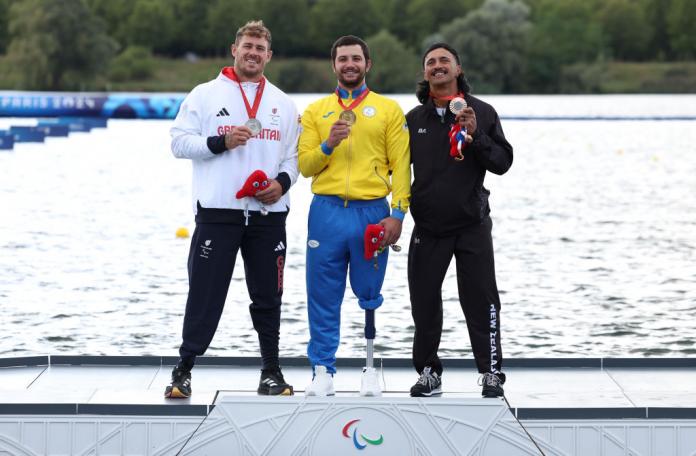 Three men stand on a podium next to a river, holding up medals