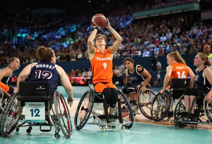 A wheelchair basketball player in a Team Netherlands uniform prepares to take a shot, with players in Team USA uniforms surrounding her