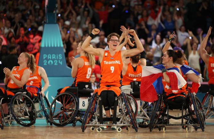 The Dutch women's wheelchair basketball team clap and raise their arms in the air, with one woman in the middle lifting up both arms and smiling