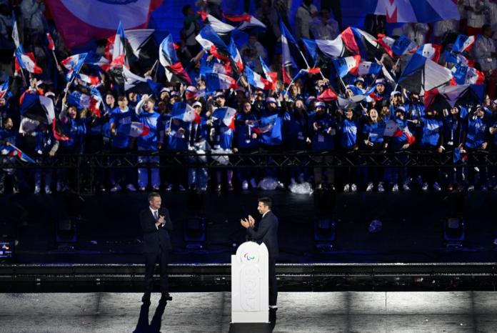 Two men in dark suits address a huge crowd with French flags in the background