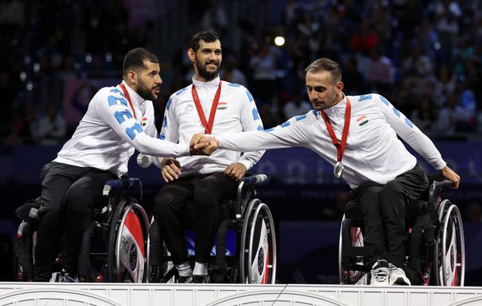 Three male wheelchair fencing athletes are celebrating on the podium after receiving silver medals