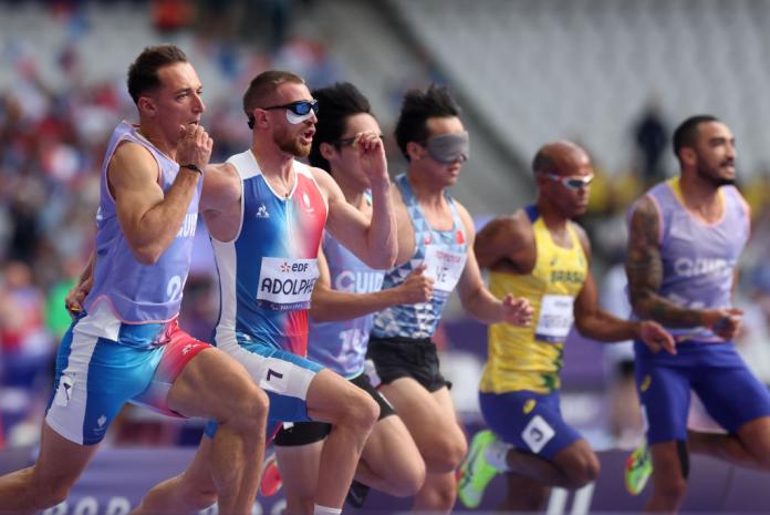 Three male sprinters and their guides are competing. The athletes are wearing blackout goggles and they are running next to their guides