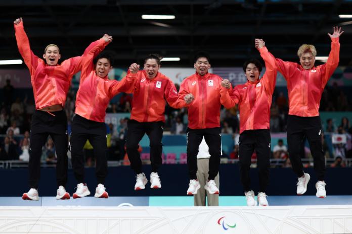 Six male goalball athletes hold hands and jump on the podium