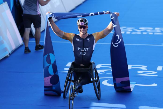 A male athlete in a racing wheelchair celebrates his victory by holding the finish tape above his head