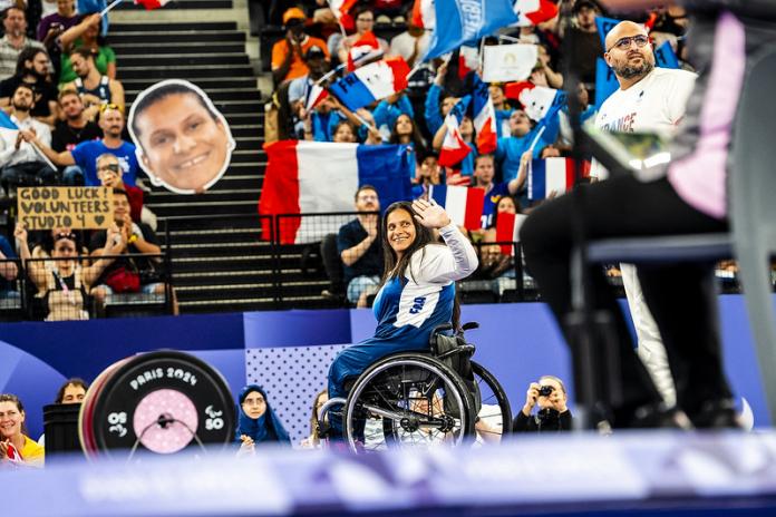A female Para powerlifter, who is using a wheelchair, waves at the crowd. The venue is packed with spectators holding French flags.