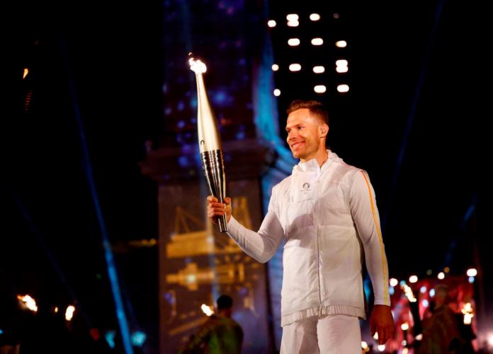 Markus Rehm, atleta masculino de salto de longitud, viste un uniforme blanco y sostiene la antorcha de París 2024 durante la Ceremonia de Apertura de los Juegos Paralímpicos París 2024
