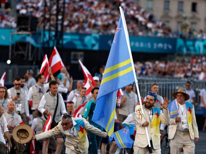 Un atleta masculino con cabello azul y gafas de sol azules lleva la bandera de Aruba durante la Ceremonia de Apertura.