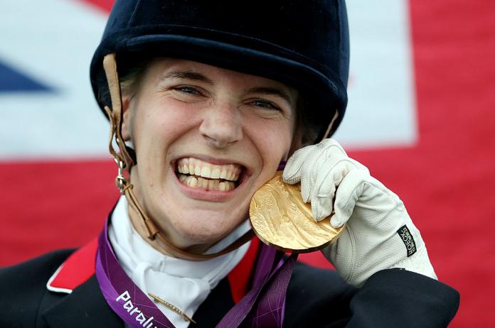 A female Para equestrian athlete smiles. She is holding a gold medal with her left hand