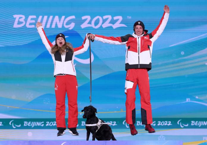 A female and male athletes jump on the podium after receiving their gold medals. A black guide dog is also on the podium.