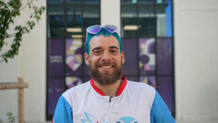 Un atleta masculino con pelo azul y gafas de sol en forma de corazón sonríe a la cámara