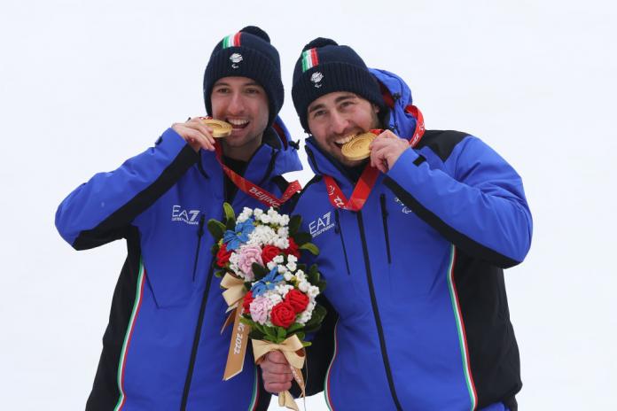 Two male athletes wearing blue winter jackets are posing for a photo. They are biting their gold medals