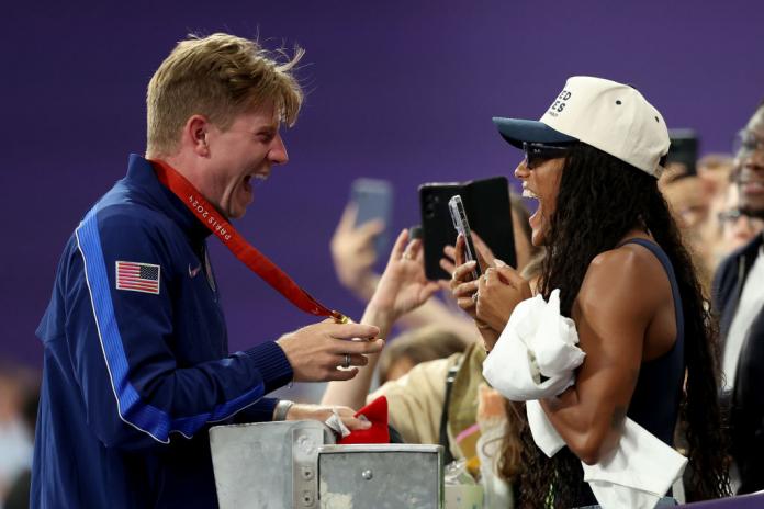 Un atleta masculino muestra su medalla de oro a una mujer en las gradas. Ella sostiene su teléfono inteligente