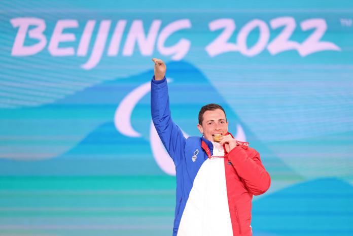 A male athlete wearing a blue, white and red jacket smiles on the podium