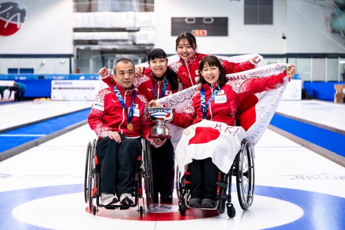 A male and female wheelchair curling athletes pose for a photo with two female officials. They are holding a trophy.
