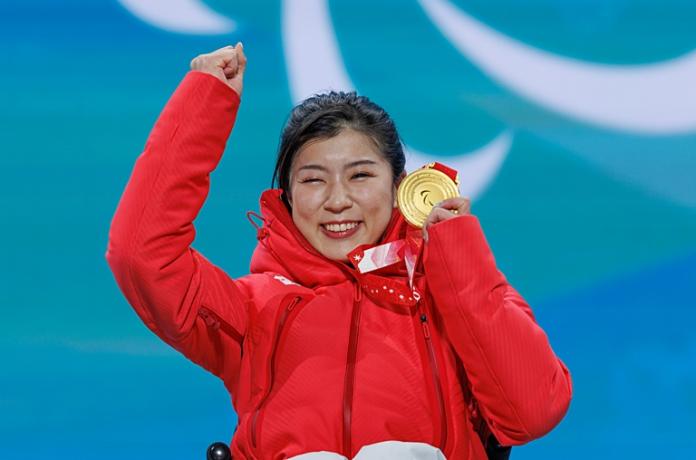 A female athlete wearing a red winter jacket is posing for a photo after receiving a gold medal