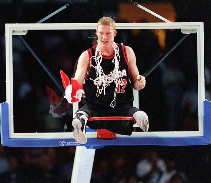 A male wheelchair basketball player is celebrating after a victory. He is sitting on the basketball backstop, with the net around his neck