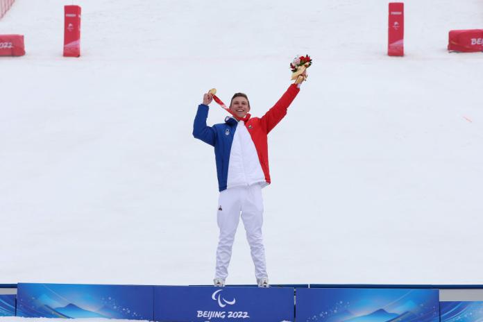 A male athlete posing on the podium. He is raising his hands and holding a flower bouquet with his left hand and a gold medal with his right hand