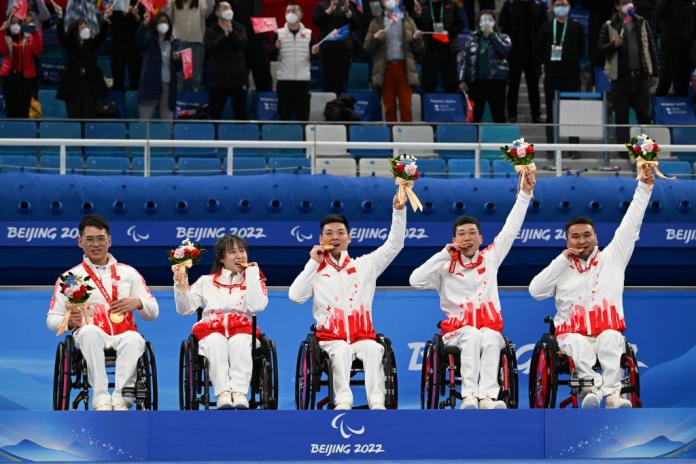 Five wheelchair curling athletes pose for a photo on the podium after receiving their gold medals and bouquets. 