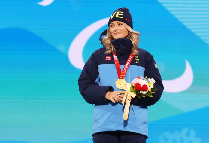 A female athlete wearing a blue winter jacket smiles on the podium after receiving a gold medal at the Beijing 2022 Paralympics.