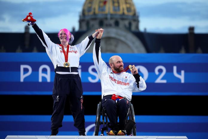 A male athlete in a wheelchair and a female athlete who has pink hair is raising their hands to celebrate after receiving their Paralympic gold medals
