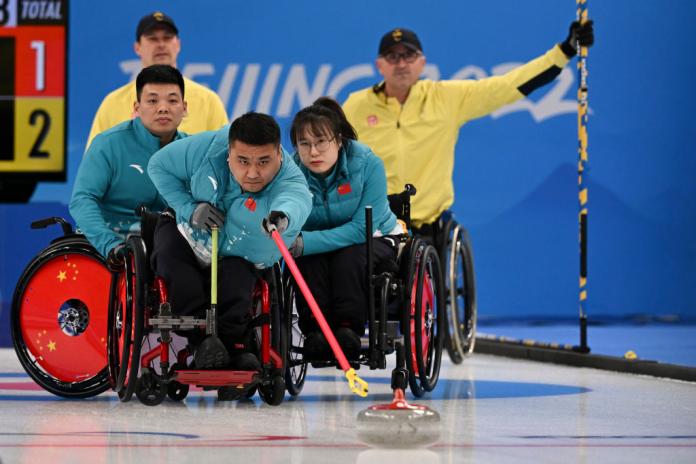 A male wheelchair curling athlete in action. He is trying to reach a curling stone with a red stick while his teammates are holding onto his wheelchair