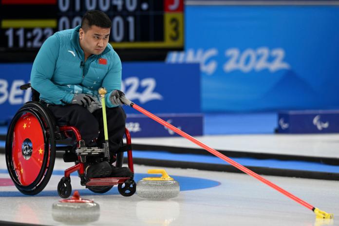 A male wheelchair curling athlete is in action. He is holding a long red stick on the ice