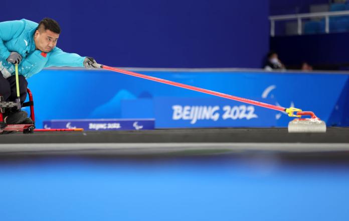 A male wheelchair curling athlete reaching for a curling stone with a red stick