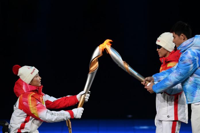 Two athletes wearing winter jackets hold Paralympic torches and pass the Paralympic flame during the Beijing 2022 Opening Ceremony