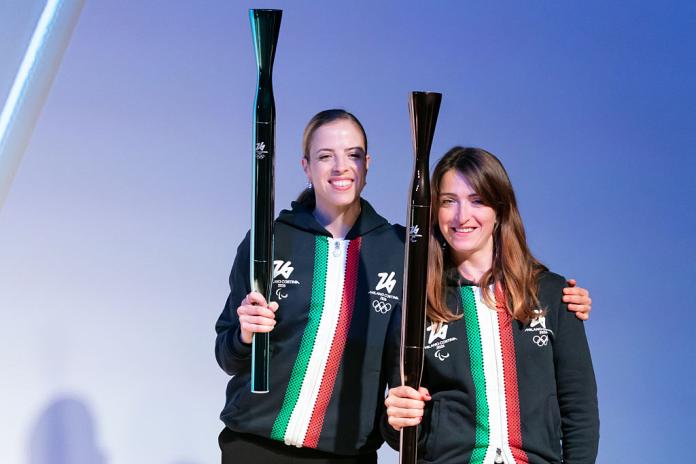 Carolina Kostner and Martina Caironi are posing for a photo while holding the torches of the Olympics and Paralympics