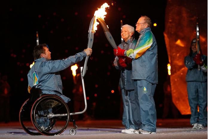 Three torchbearers pass the flame during the Vancouver 2010 Opening Ceremony. A man in a wheelchair has a device attached on the wheel that is holding the torch.