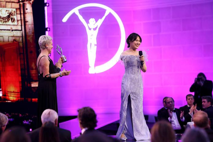 A female Para athlete wearing a silver dress is speaking on stage. There are two people presenting her the award