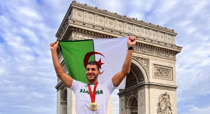 A male Para canoe athlete holds the Algerian flag in front of the Arch de Triomphe in Paris.