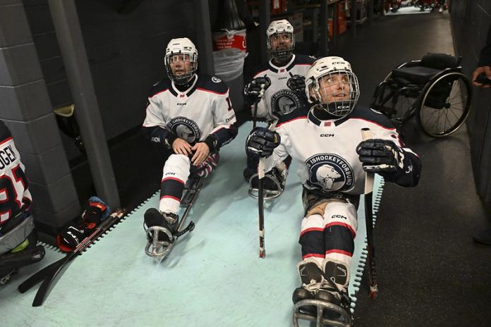Three Para ice hockey players are in their sleds and on plastic ice at an indoor venue