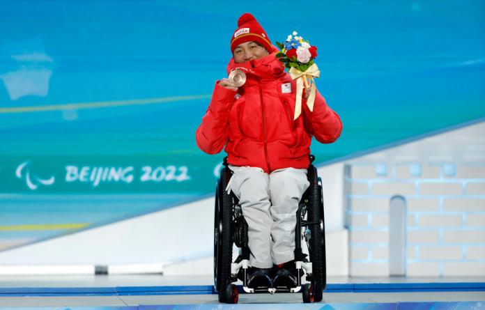 A male Paralympic athlete, who uses a wheelchair, is posing for a photo after receiving a bronze medal at Beijing 2022