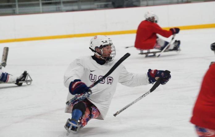 A male Para ice hockey player, wearing a helmet and a white Team USA gear, is in action on the ice. He is holding two sticks.