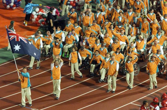 Around 50 people wearing orange vests are parading behind a male athlete waving the Australian flag