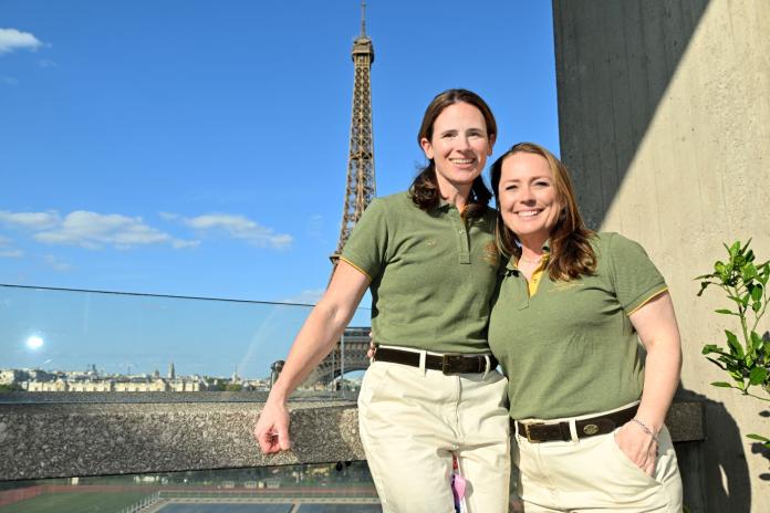 Two female officials wearing green polo shirts and khaki pants pose in front of the Eiffel Tower