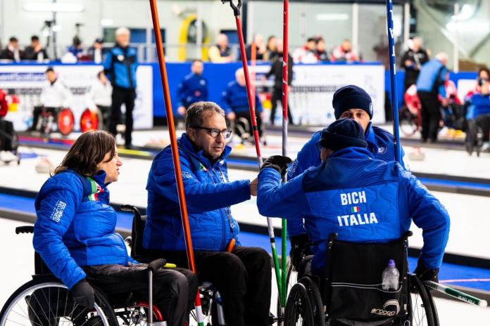 Wheelchair curling athletes, wearing a blue winter jacket, are doing a fist bump 