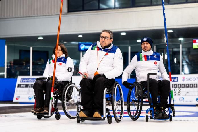 Three wheelchair curling athletes wearing white Italian uniforms are on the ice