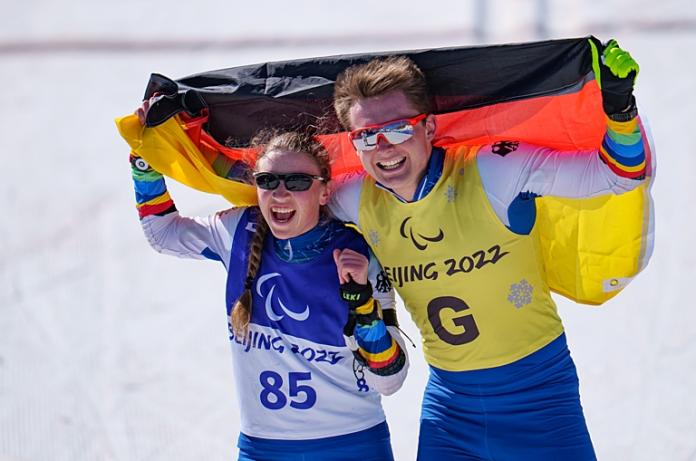 A female Paralympic skier and a sighted male guide are celebrating by holding the German flag