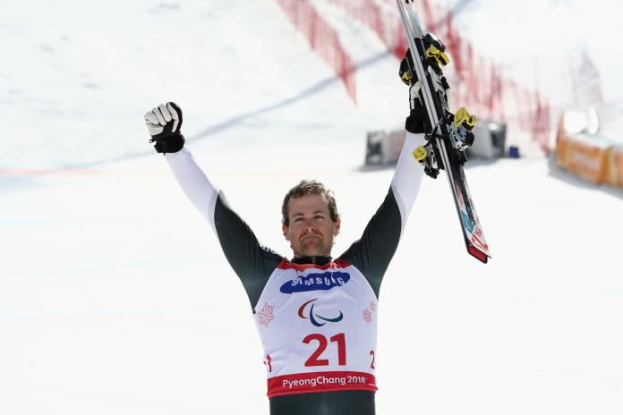 A male Para alpine skier celebrating winning with his arms wide open, positioned in the sky, while he holds his skis 