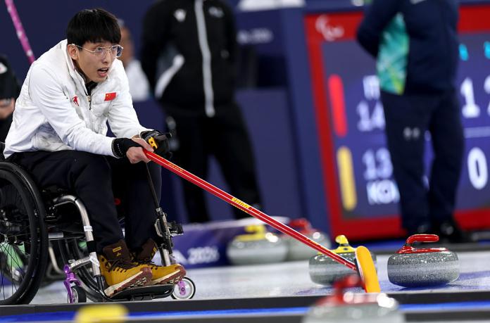 A male wheelchair curling athlete is holding a red stick and places it between two curling stones 