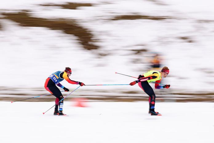 Two skiers are in action, one athlete is holding onto the poles of the other