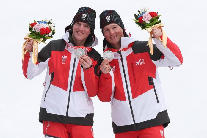 One male Para alpine skier posing alongside his guide, celebrating his silver medal, while holding flowers