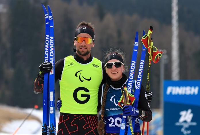 A female Para cross country skier smiling at the camera with her guide, while both of them are holding their skis