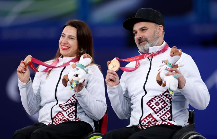 One female and one male wheelchair curler posing for the picture while holding their bronze medal and smiling